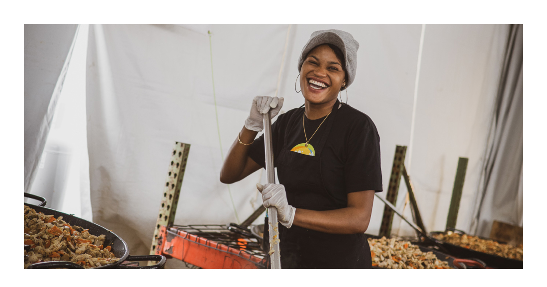 Image description: Woman smiling at the camera and holding a long ladle next to large pans of food.